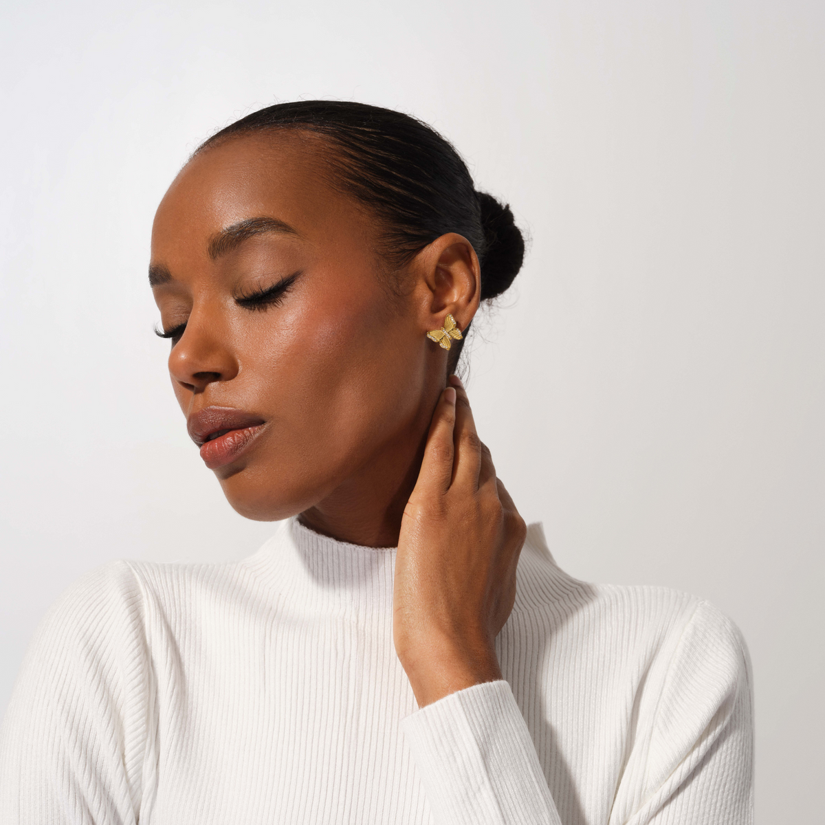 A woman with her hair in a bun wearing a white ribbed mock neck top and a small gold geometric stud earring, posing against a light gray studio background.