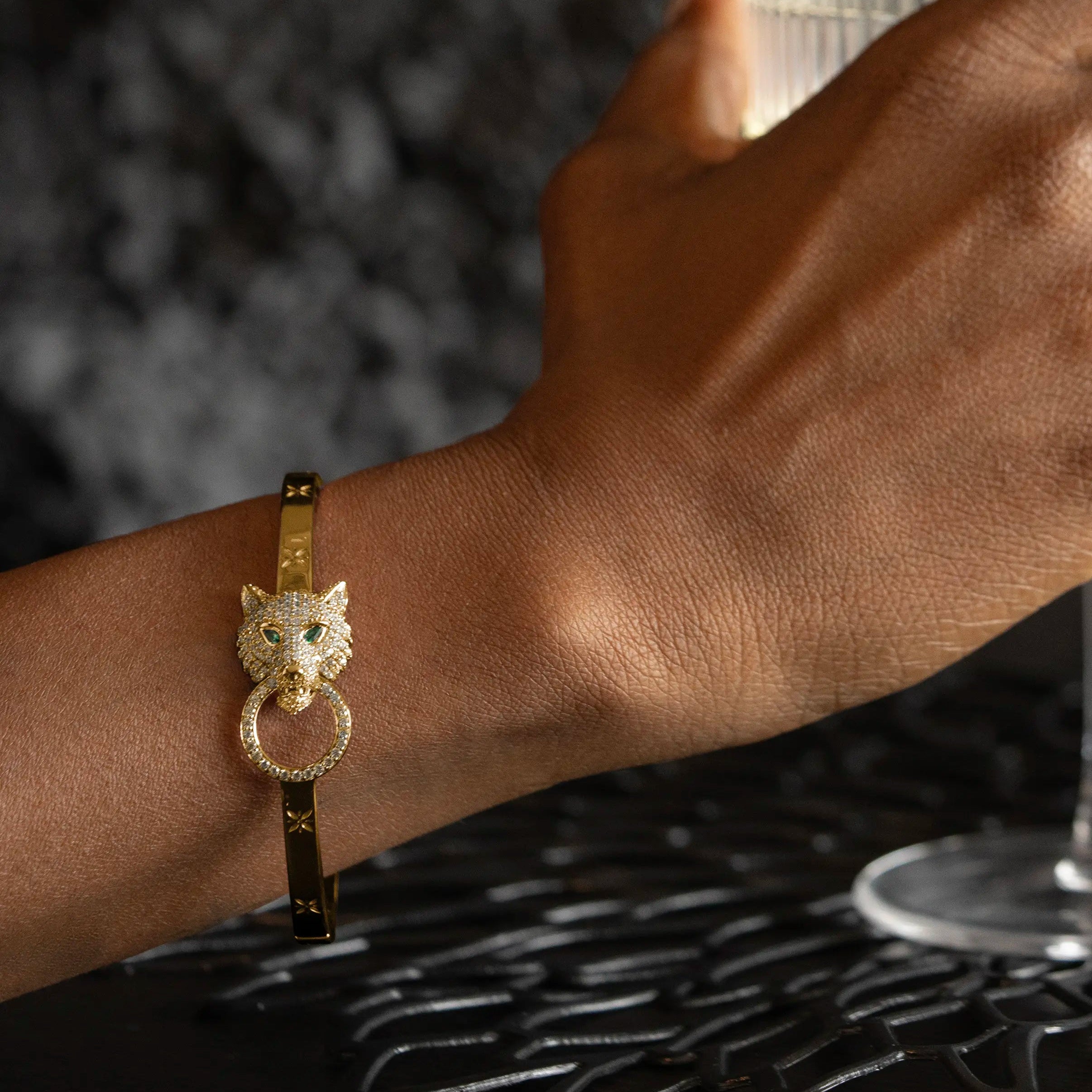 Hand holding a glass with a gold ring and bracelet on a textured black surface